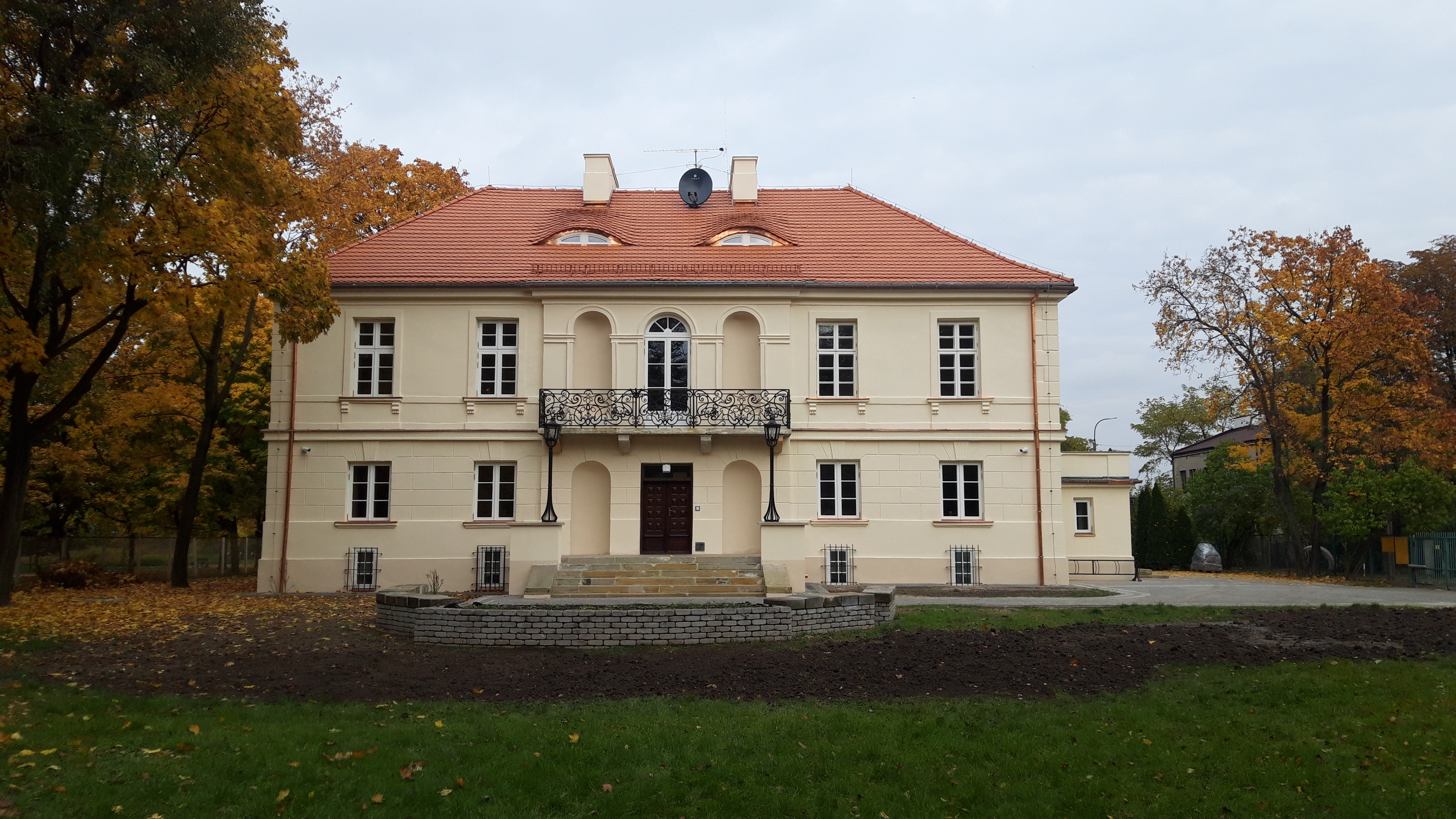 Zdjęcie przedstawia zabytkowy budynek szkoły, dziewiętnastowieczny dworek, po remoncie. Budynek ma kolor brzoskwiniowy, dach koloru ceglastego. Stoi w otoczeniu parku. Pora roku na zdjęciu to jesień.
