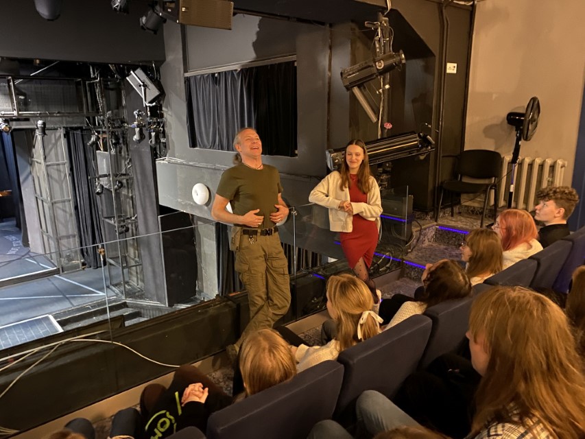 Zdjęcie kolorowe. Grupa uczniów siedzi na widowni teatru i słucha mówiącego aktora.