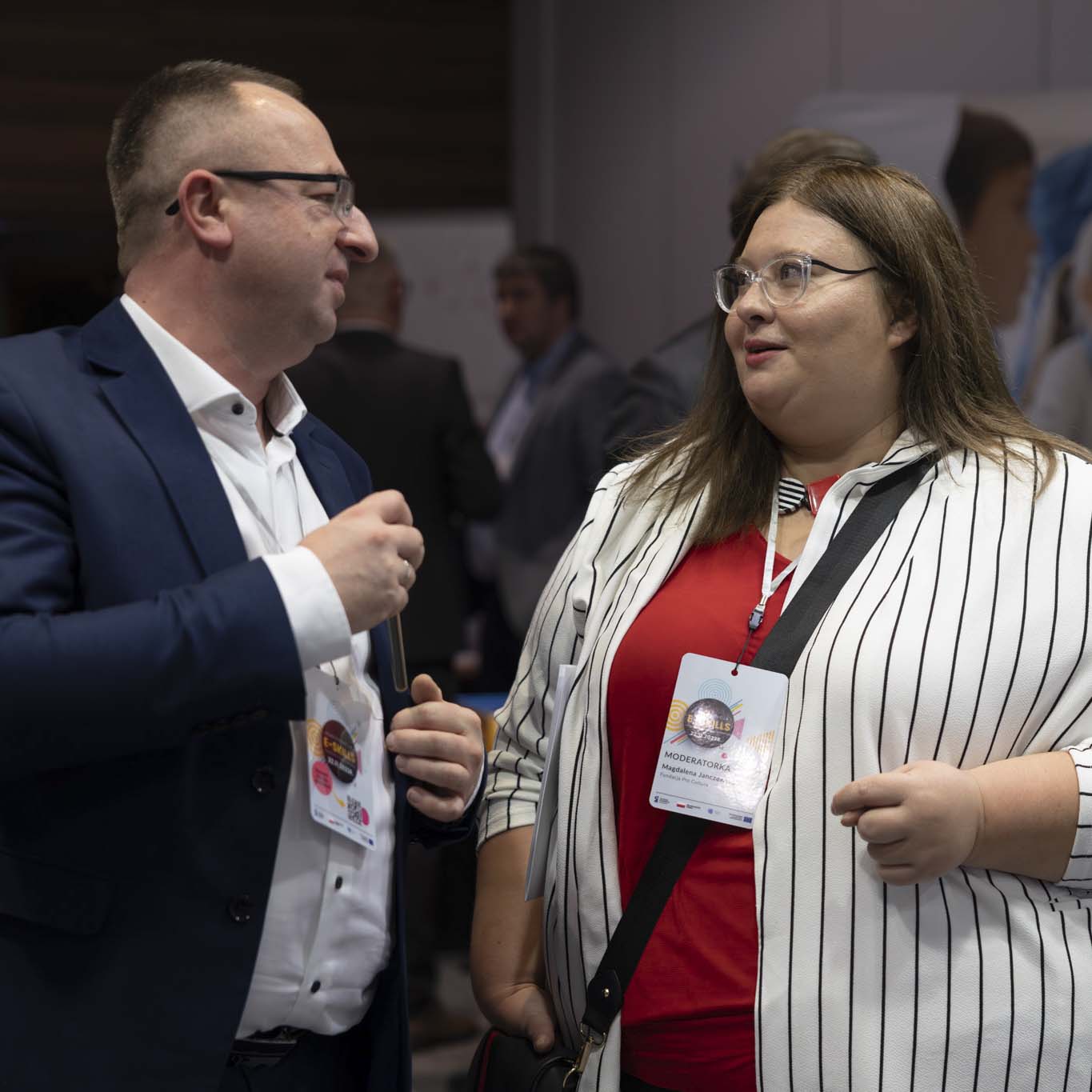 Kobieta i mężczyzna rozmawiają, patrzą na siebie. W tle pozostali uczestnicy konferencji.