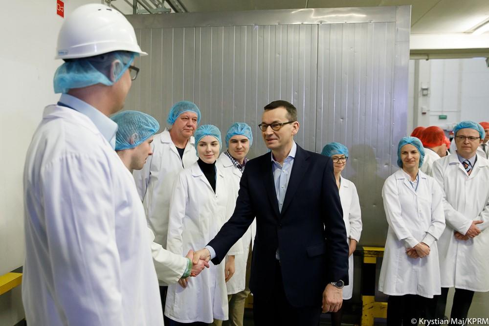 Premier Mateusz Morawiecki podaje rękę jednemu z pracowników zakładu. Obok pozostali pracownicy zakładu stoją ubrani w białe fartuchy i niebieskie siatki na włosy.