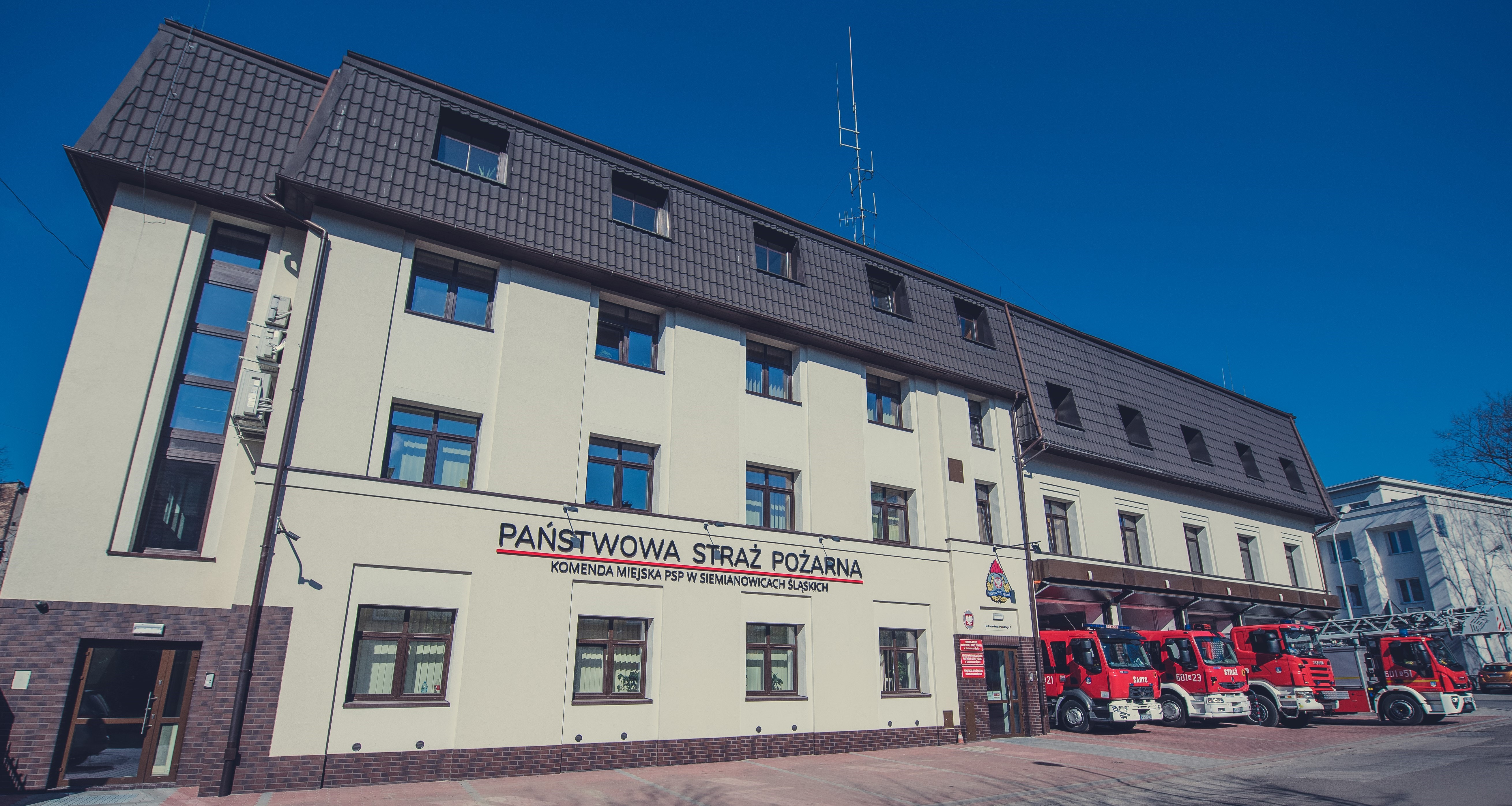 Na zdjęciu widoczny jest budynek straży pożarnej. Na fasadzie widnieje napis: "Państwowa Straż Pożarna", co oznacza, że jest to siedziba jednostki zawodowej straży pożarnej w Siemianowicach Śląskich. Budynek jest trzypiętrowy, w jasnych kolorach, z ciemnym dachem i licznymi oknami. Przed budynkiem zaparkowane są cztery czerwone wozy strażackie.