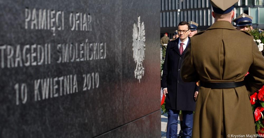 Premier Mateusz Morawiecki stoi za pomnikiem pamięci ofiar tragedii smoleńskiej, a obok stoi żołnierz.