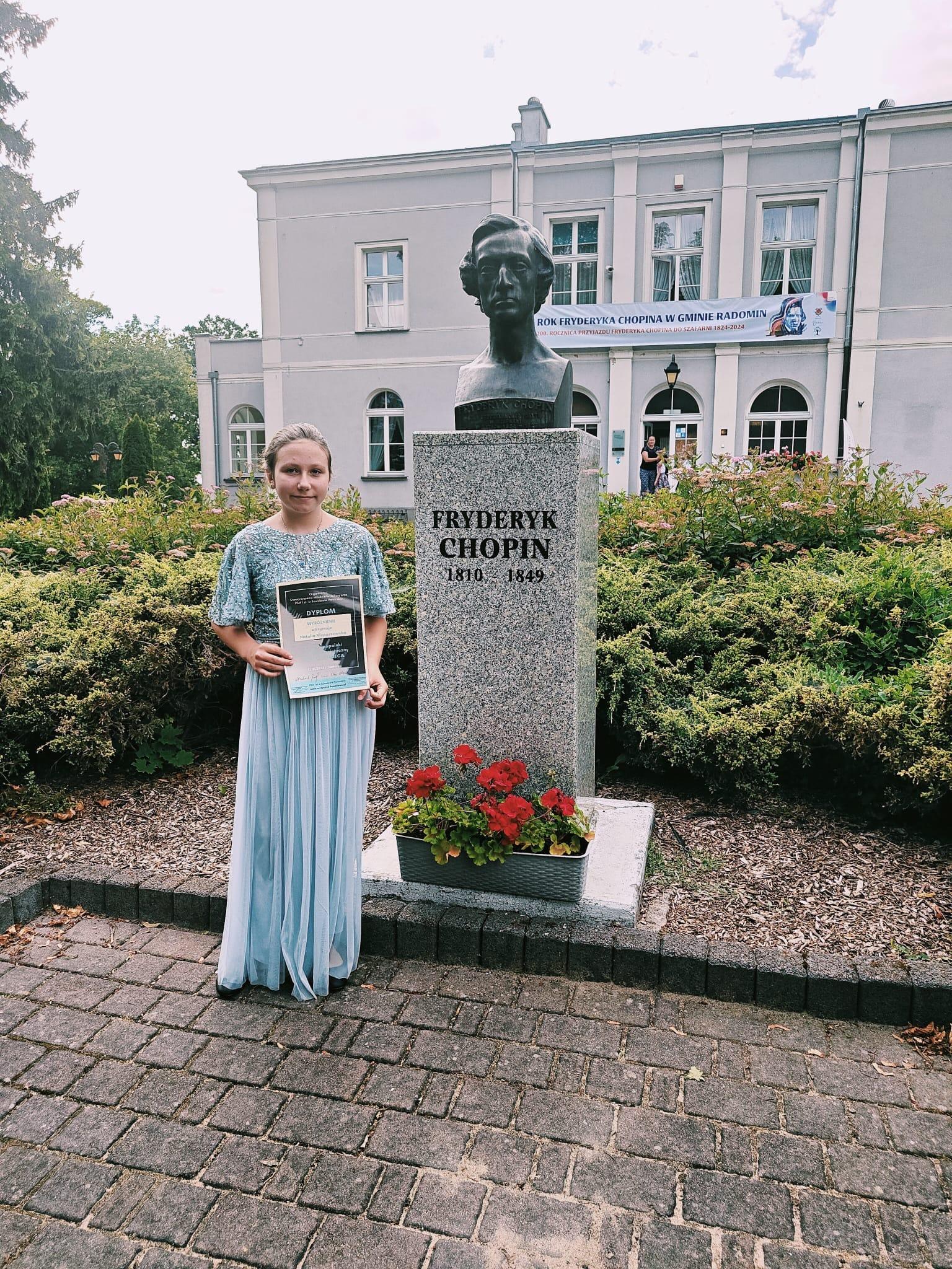 Na zdjęciu znajduje się uśmiechnięta uczennica w długiej błękitnej sukience, w rękach trzyma dyplom konkursowy. Stoi przy pomniku szkolnym Fryderyka Chopina, który znajduje się przed budynkiem szkolnym .