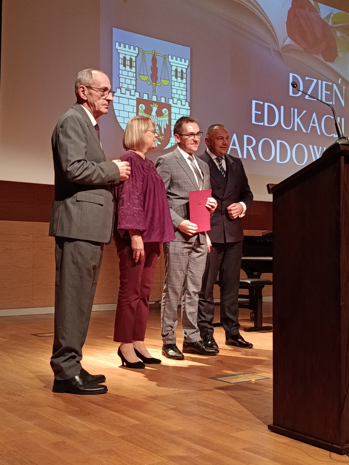 Czworo dorosłych osób stoi na scenie w oświetlonej sali. Scena ma drewnianą podłogę. Po lewej stronie ekranu za nimi znajduje się herb miasta wyświetlany na dużym ekranie. Po prawej stronie ekranu wyświetlany jest tekst „DZIEŃ EDUKACJI NARODOWEJ” i otwarta książka. Po prawej stronie stoi mównica. Z lewej strony stoi mężczyzna w szarym garniturze, który trzyma rękę na ramieniu stojącej obok kobiety. Kobieta ma na sobie fioletową bluzkę i bordowe spodnie. Po prawej stronie kobiety stoi mężczyzna w szarym, wzorzystym garniturze, który trzyma w dłoni czerwony dokument, prawdopodobnie nagrodę. Obok niego, najbardziej po prawej, stoi mężczyzna w ciemnym garniturze i krawacie. W tle widoczny jest fragment fortepianu