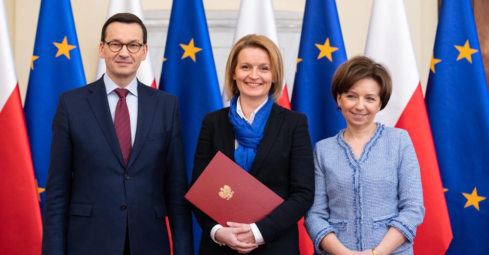 Premier Mateusz Morawiecki, Barbara Socha i minister Marlena Maląg na tle flag.