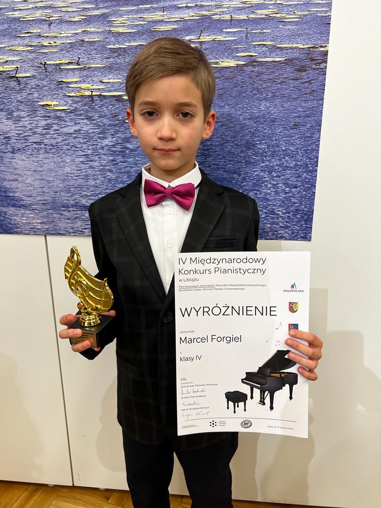 Zdjęcie przedstawia ucznia ubranego w ciemny garnitur, który trzyma statuetkę oraz dyplom konkursowy.