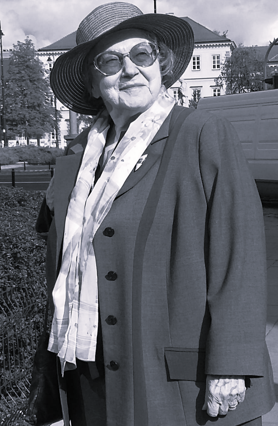 Czarnobiałe zdjęcie przedstawiające na tle budynku kobietę w okularach przeciwsłonecznych i kapeluszu. Kobieta ubrana jest w płaszcz oraz szalik. 