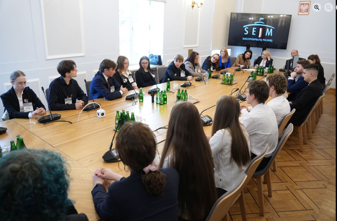 Zdjęcie przedstawia oficjalne spotkanie odbywające się w jasnej, klasycznie urządzonej sali konferencyjnej w Sejmie Rzeczypospolitej Polskiej. Liczna grupa młodych ludzi oraz kilka dorosłych osób siedzi wokół długiego, drewnianego stołu ustawionego w kształcie podkowy. Uczestnicy są ubrani w stroje galowe, a wielu z nich ma przypięte identyfikatory. Na stole, przed poszczególnymi osobami, znajdują się mikrofony konferencyjne, szklanki oraz liczne zielone butelki z wodą mineralną. W głębi pomieszczenia, po prawej stronie, na ścianie wisi duży monitor wyświetlający logo Sejmu Rzeczypospolitej Polskiej na czarnym tle, a obok niego widoczny jest godło Polski. Sala ma wysokie, jasne ściany zdobione subtelnymi sztukateriami oraz parkiet ułożony w klasyczną jodełkę. Przez duże okno w tle wpada naturalne światło, które rozjaśnia całą scenę. Panuje atmosfera skupienia i powagi, sugerująca przebieg oficjalnych obrad lub konsultacji społecznych.