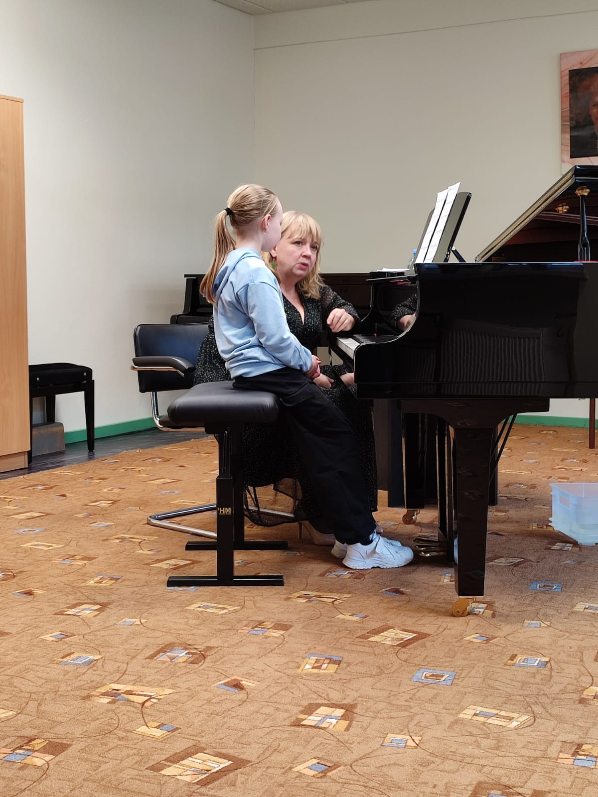 Uczennica siedząca przy fortepianie, obok niej nauczycielka. Po lewej stronie widoczna szafa. Na ścianie wisi portret Ignacego Jana Paderewskiego. 