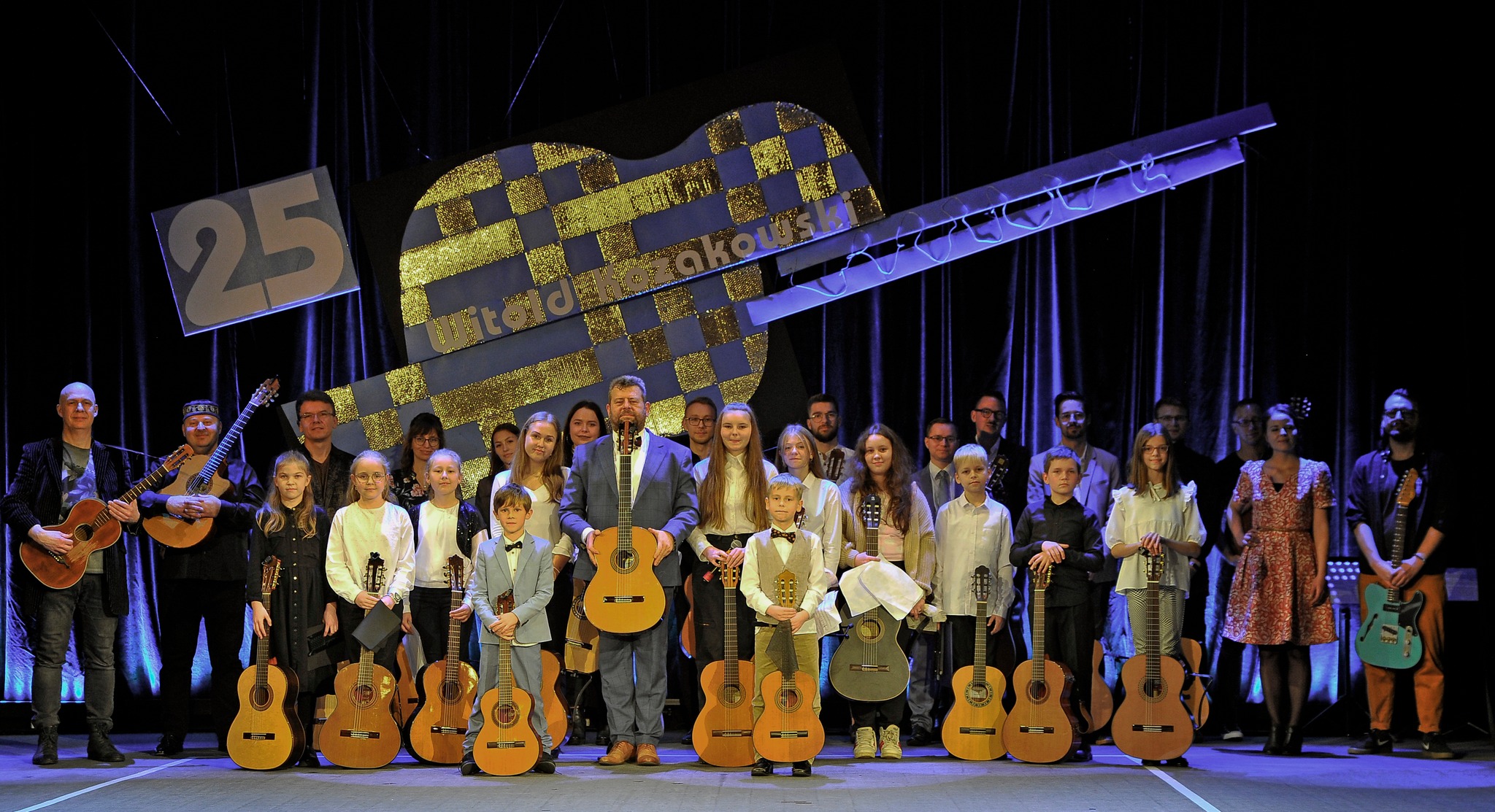 Zdjęcie grupowe wykonane na scenie w Kłodzkim Ośrodku Kultury, wszystkie osoby na zdjęciu trzymają gitarę, a w tle widoczna jest grafika gitary z napisem 25 Witold Kozakowski