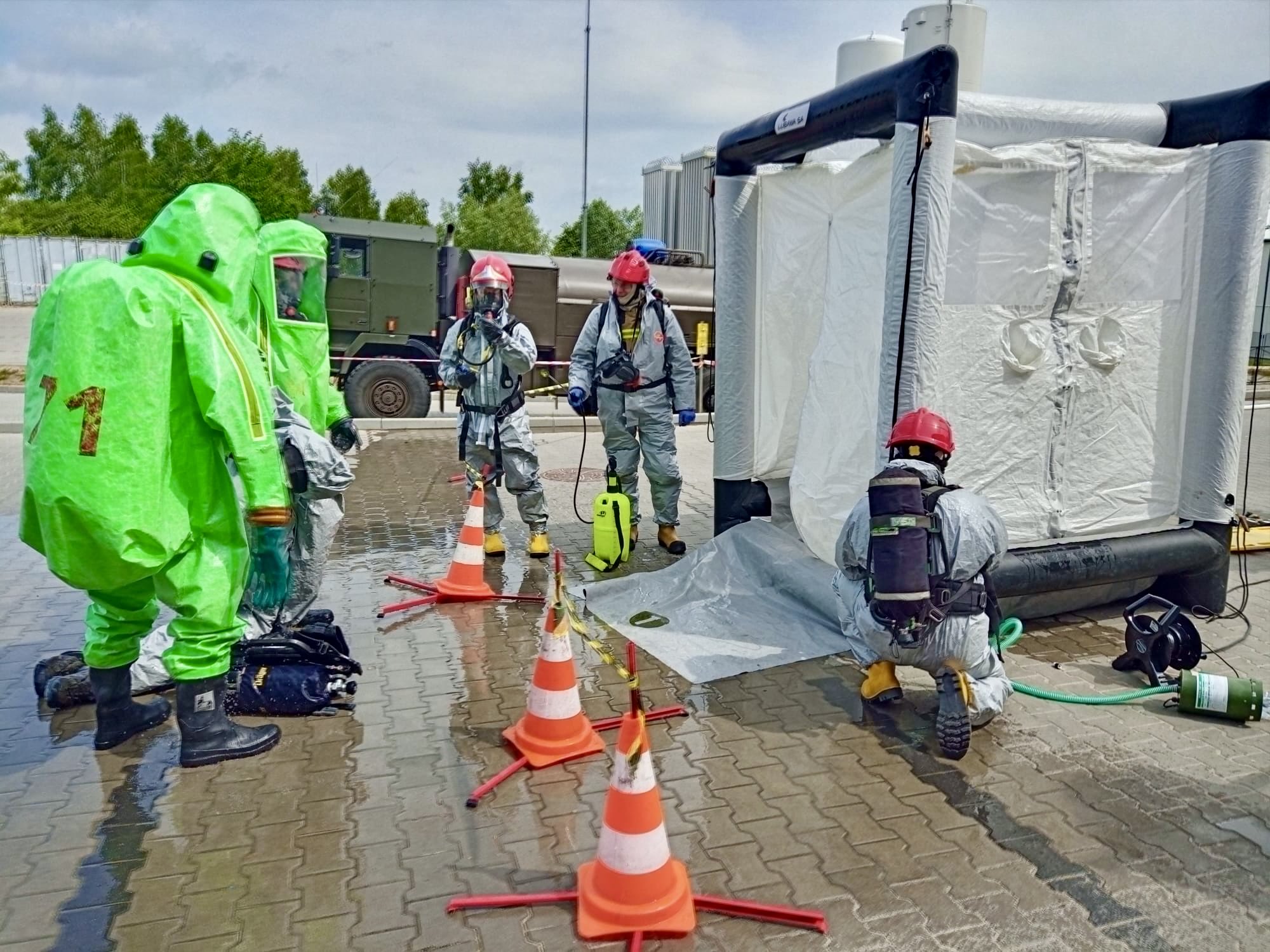 Strażacy ratownictwa chemicznego w skafandrach na placu zewnętrznym podczas ćwiczeń, w tle samochód - cysterna