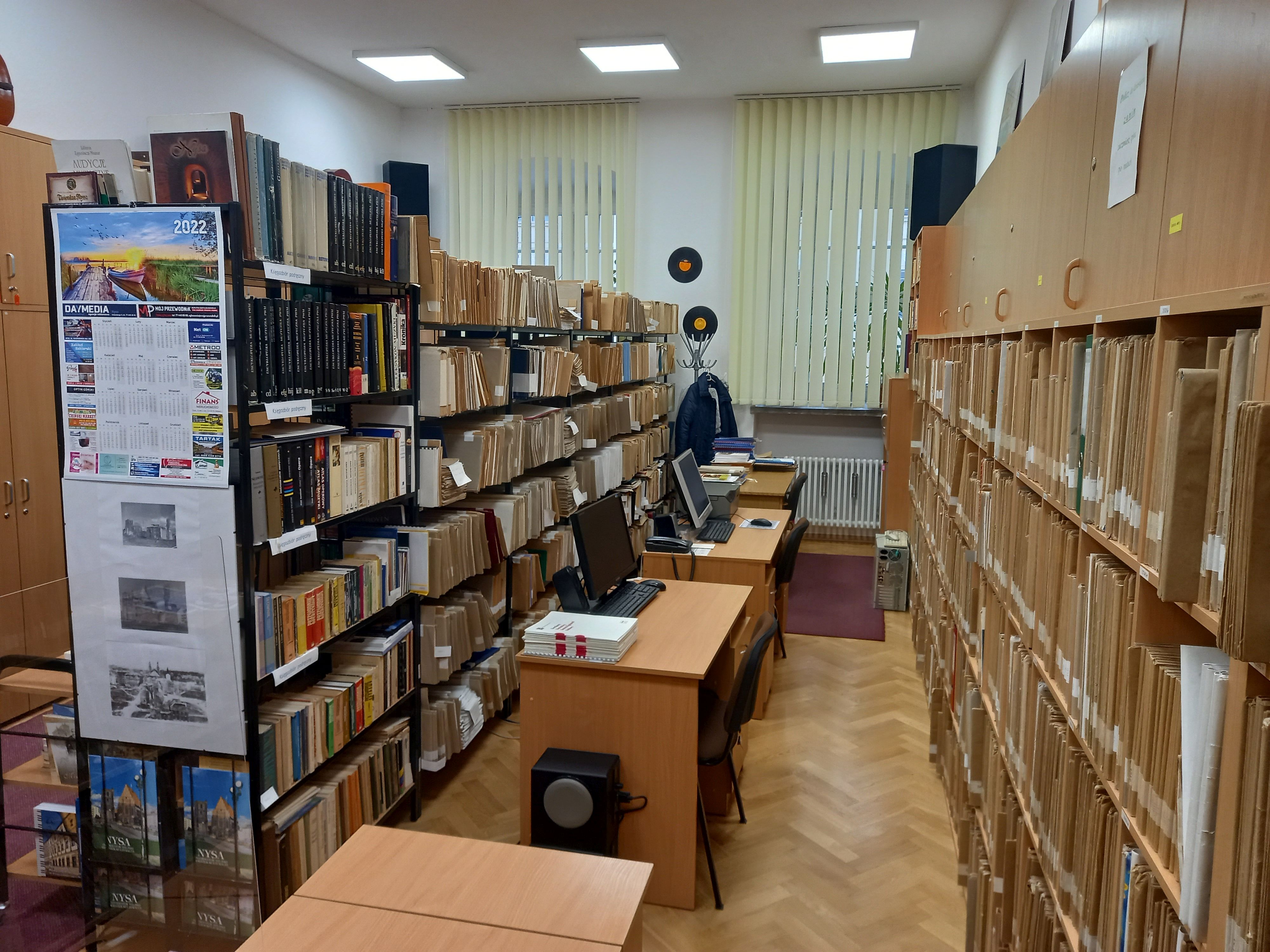 Zdjęcie przedstawia regały z nutami, książkami i szafki z płytami. w środku biurka ze stanowiskami komputerowymi.