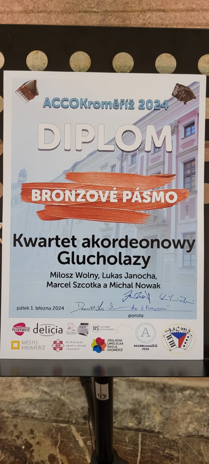 Dyplom w tle zarys klasycystycznego budynku w kolorze biało różowym prawym górnym i lewym rogu grafiki akordeonów poniżej napis w kolorze niebieskim ACCOKromeriż 2024 napis w kolorze białym dyplom poniżej na trzech pasach imitujących malowanie farbą napis brązowe pasmo kwartet akordeonowy Głuchołazy Miłosz Wolny Lukas Janocha Marcel Szczotka i Michał Nowak poniżej po czesku 1 marca 2024 piątek oraz odręczne podpisy członków jury na samym dole kolorowe logotypy organizatorów i sponsorów w języku czeskim według ustalonych znaków
