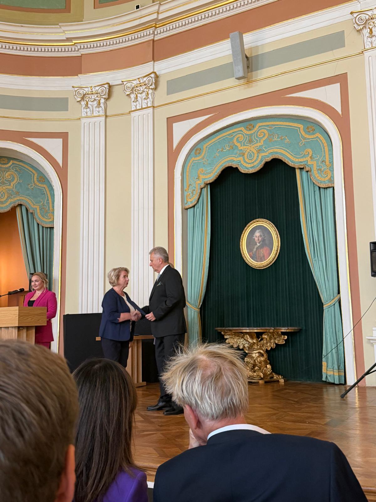 Kadr ujęty z perspektywy widowni. W centrum kobieta w ciemnym garniturze - Pani Irena Maciejewska odbiera teczkę z nominacją i potrząsa dłoń mężczyzny w czarnym garniturze - Sławomir Rogowski. Po ich lewej stronie, za jasnym, drewnianym pulpitem z mikrofonem, stoi kobieta w różowym żakiecie. Tło to bogato zdobione wnętrze z kolumnami, gzymsami i dwiema wysokimi wnękami z turkusowymi zasłonami, we wnęce po prawej stronie wisi owalny portret w złotej ramie. 