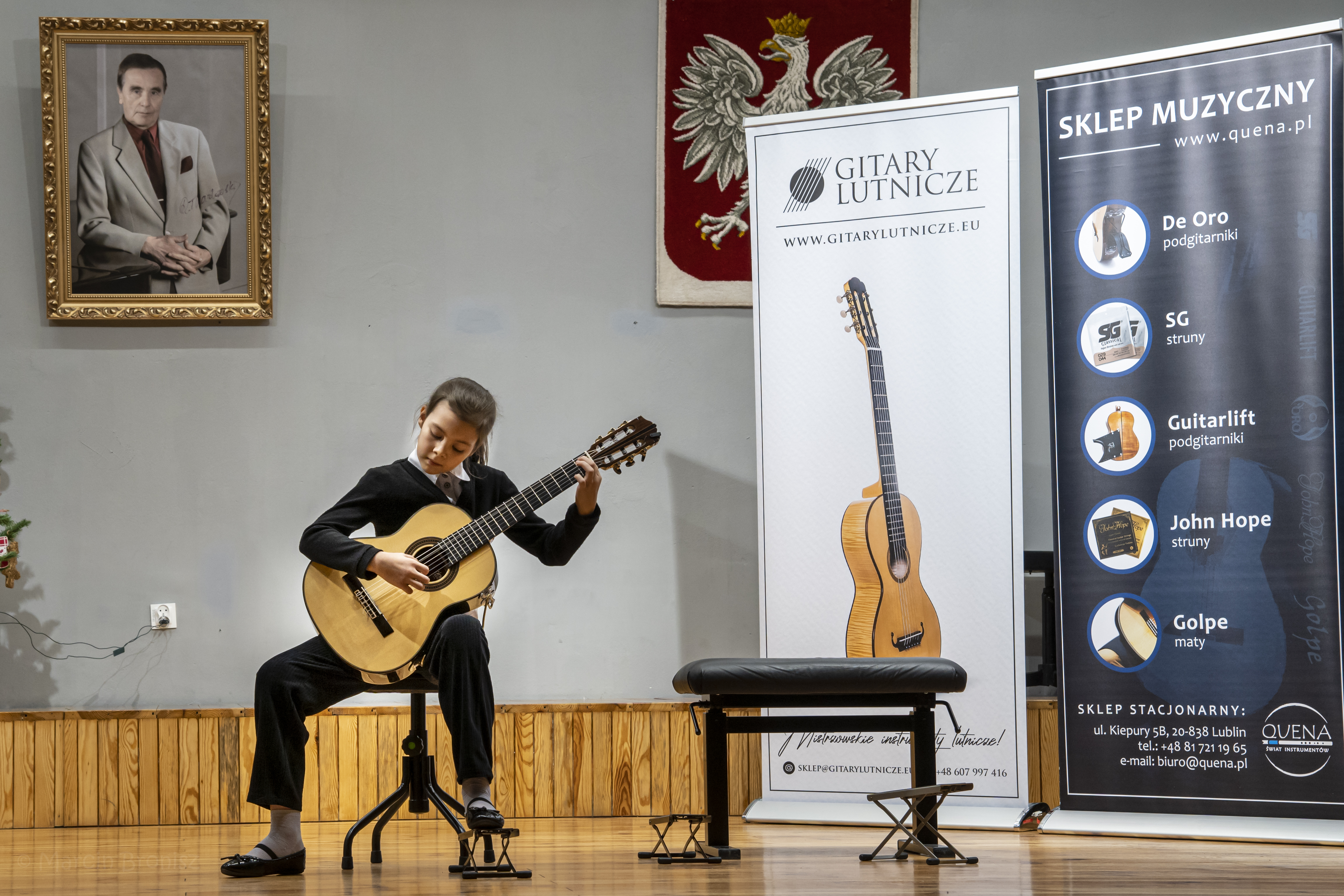 I Konkurs Gitarowy w Puławach. Sala koncertowa Państwowej Szkoły Muzycznej im. Romualda Twardowskiego w Puławach. 9.12.2023 r. Uczennica Lena Torba w czasie występu konkursowego na dużej scenie gra na gitarze. Po prawej stronie stoją dwa banery sponsora konkursu sklepu Quena. Na ścianie wisi duże godło i obraz patrona szkoły. 