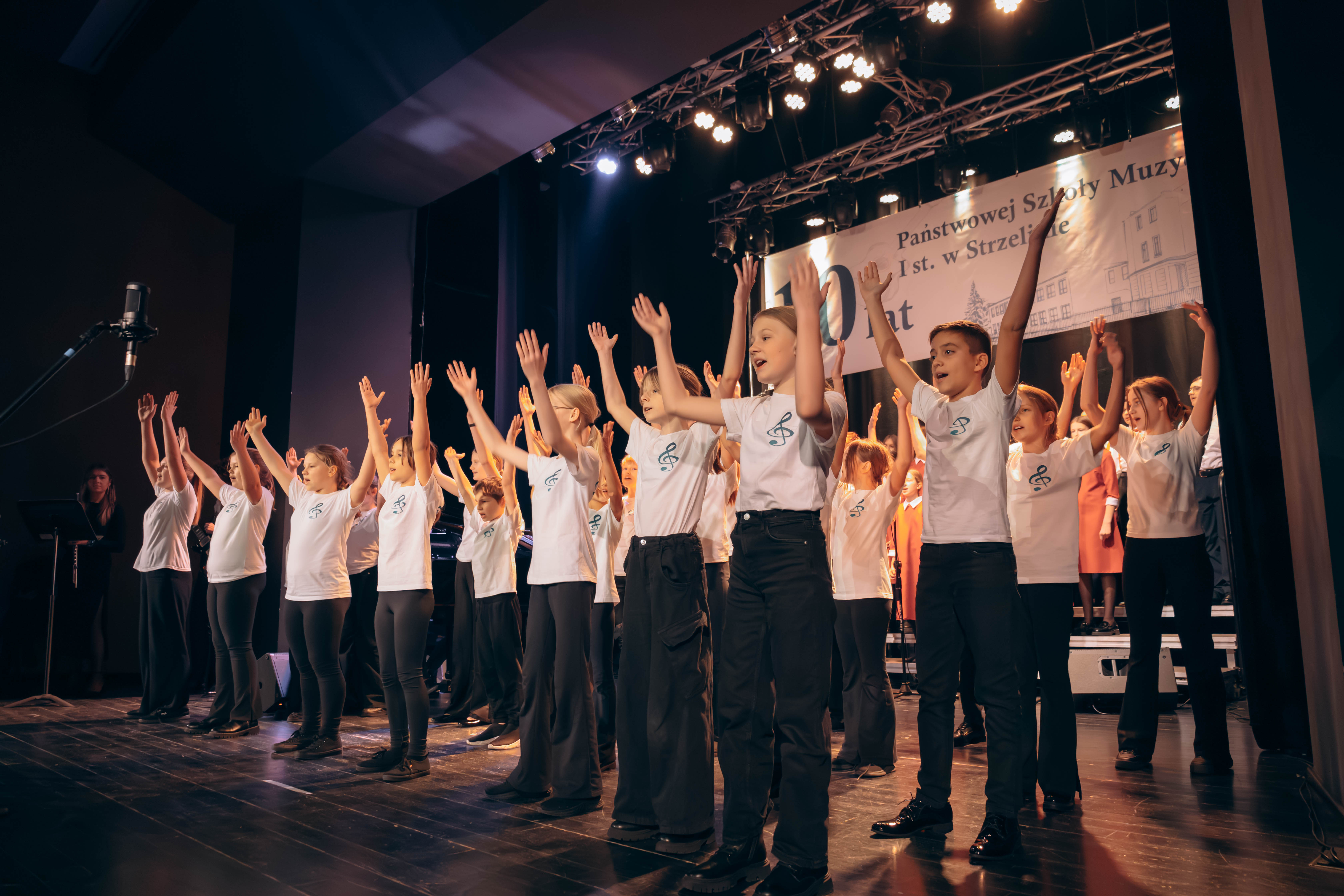 Na zdjęciu widoczna jest duża grupa dzieci ubrana w czarne spodnie i białe koszulki. Występują one na scenie.