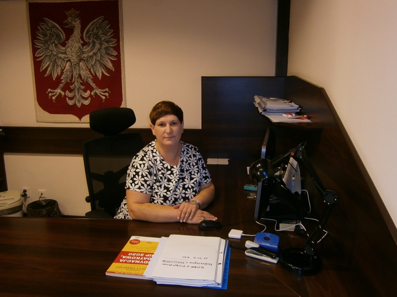 pomieszczenie, biurko za wysoką czarną ladą, wokół dokumenty i przedmioty biurowe, przy biurku siedzi kobieta