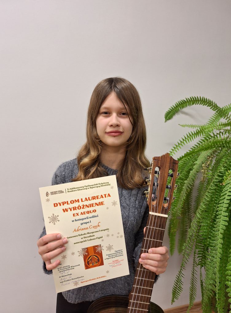 Na zdjęciu widoczna uśmiechnięta uczennica w blond rozpuszczonych włosach. W prawej ręce trzyma dyplom konkursowy, w lewej gitarę. W tle widoczna zielona roślina. 