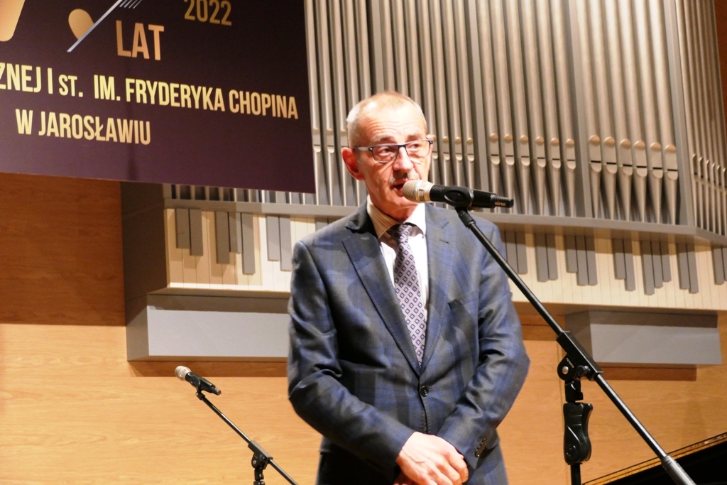Zdjęcie przedstawiające przemawiającego na scenie sali koncertowej ZPSM w Jarosławiu wizytatora CEA - pana Zenobiusza Kajdę. Po lewej stronie u góry ciemnobrązowy baner z napisem 75 lat PSM I st. im. F. Chopina w Jarosławiu,  na środku na tle prospektu w formie organów piszczałkowych z grafika klawiatury organowej poniżej, przemawiający do mikrofonu ustawionego na statywie pan wizytator