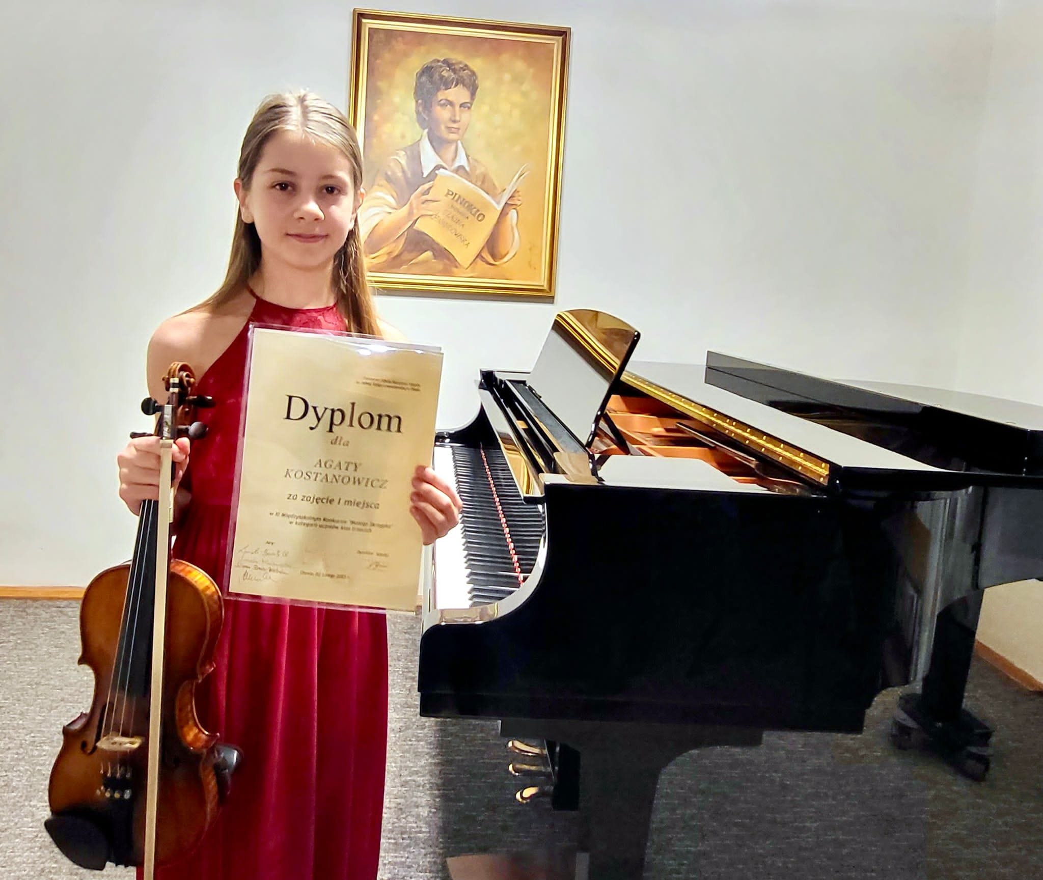 Na zdjęciu widoczna jest uśmiechnięta uczennica w czerwonej sukni trzymająca skrzypce oraz dyplom konkursowy. Za nią stoi czarny fortepian.