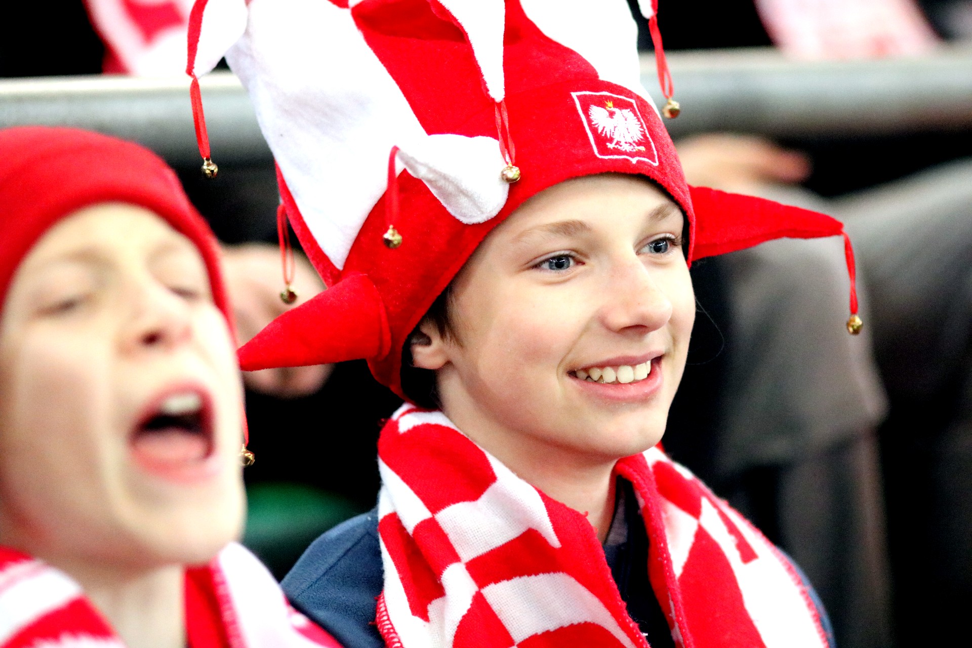 Dwoje dzieci w biało-czerwonych szalikach, jeden chłopiec ma na głowie wysoką biało-czerwoną rogatą czapkę z dzwoneczkami i orłem.