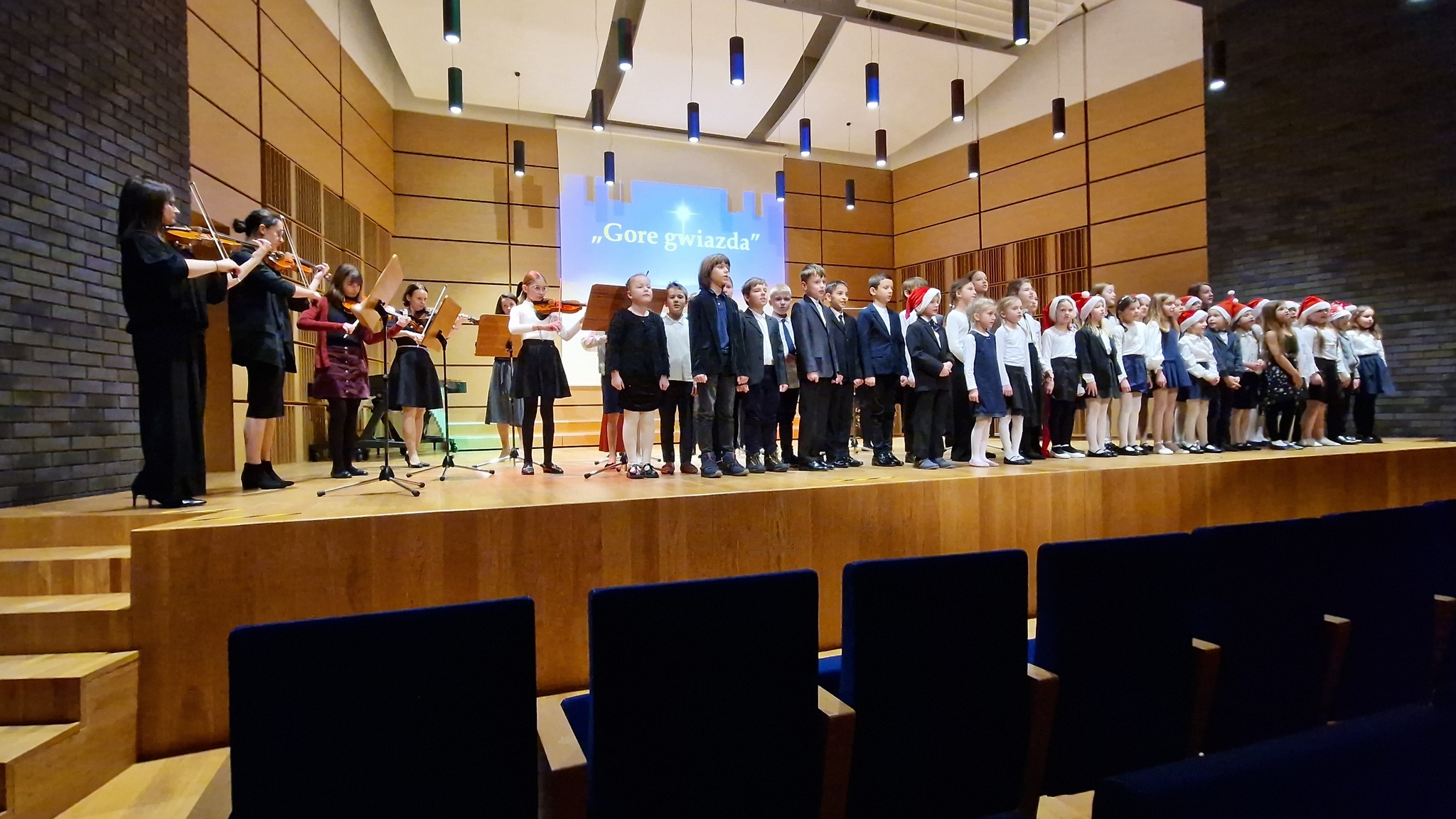 Uczniowie śpiewający i grający na scenie. Przed osobami grającymi stoją pulpity z nutami. Na dole zdjęcia widoczna jest widownia.