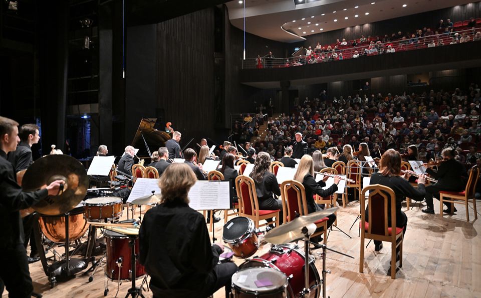 Zdjęcie. Na estradzie Sali Widowiskowej Nyskiego Domu Kultury występuje orkiestra, widok od kulis. Na pierwszym planie orkiestra, w tle widać wypełnioną "po brzegi" widownię.