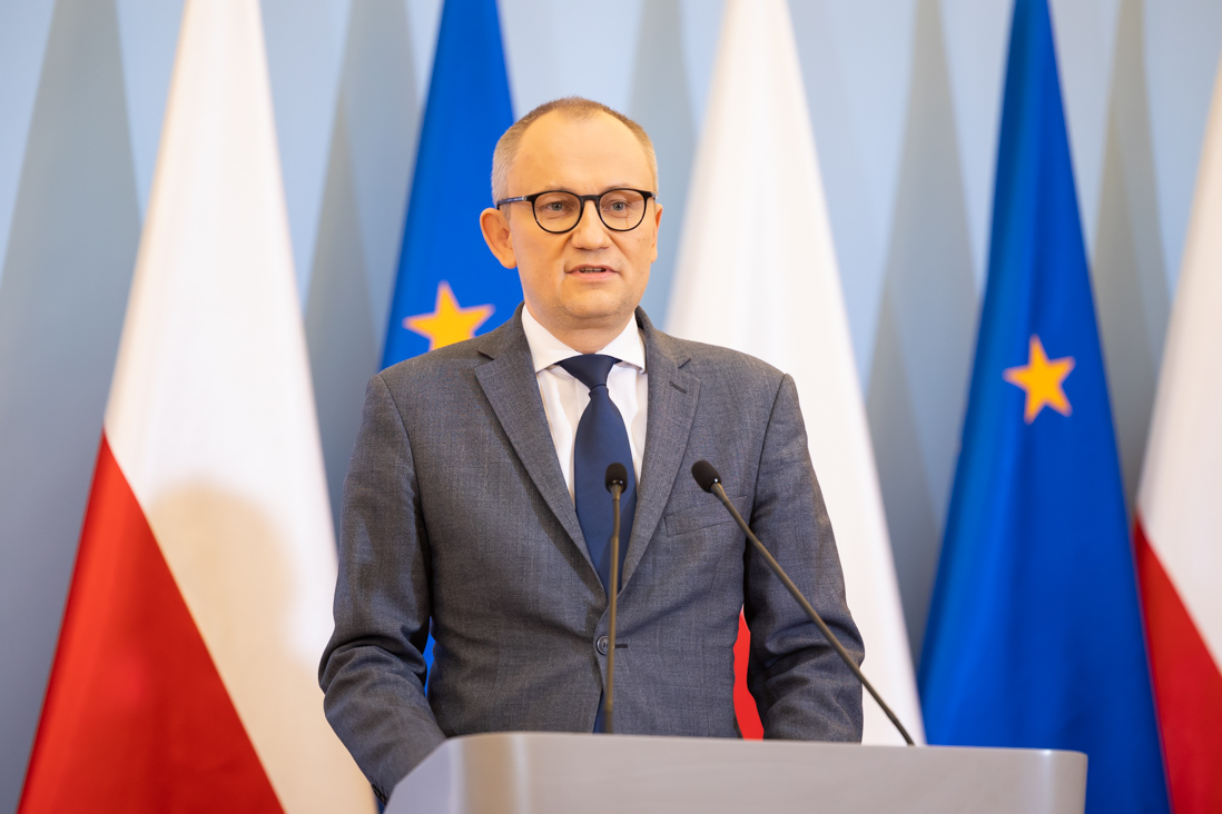 Wiceminister Błażej Poboży podczas konferencji prasowej. W tle flagi Polski i UE