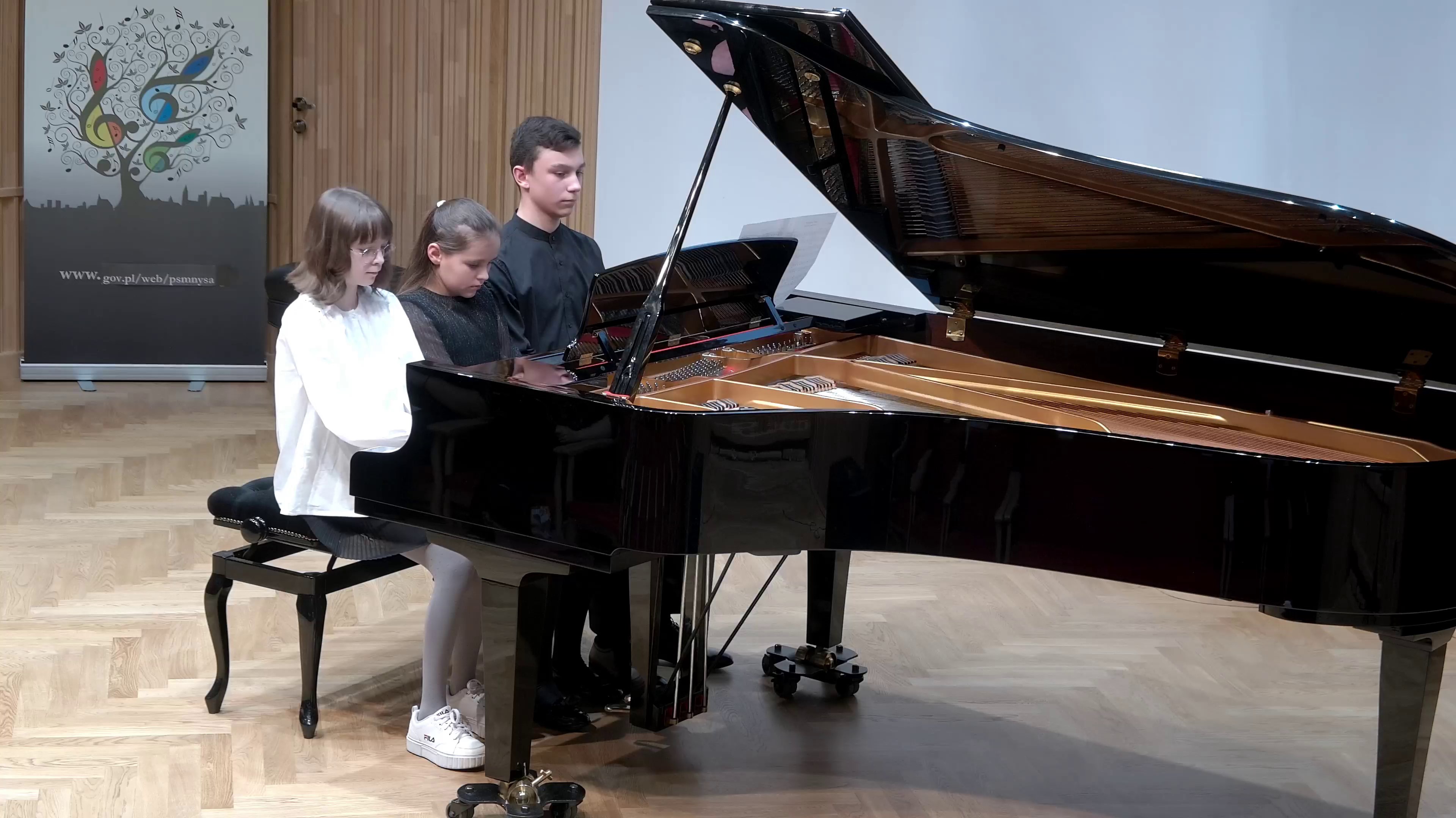 Zdjęcie. Na estradzie sali koncertowej troje uczniów gra utwór na sześć rąk na dużym czarnym fortepianie. W tle z lewej strony baner PSM Nysa.