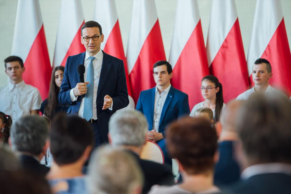 Premier Mateusz Morawiecki na tle flag biało-czerwonych.