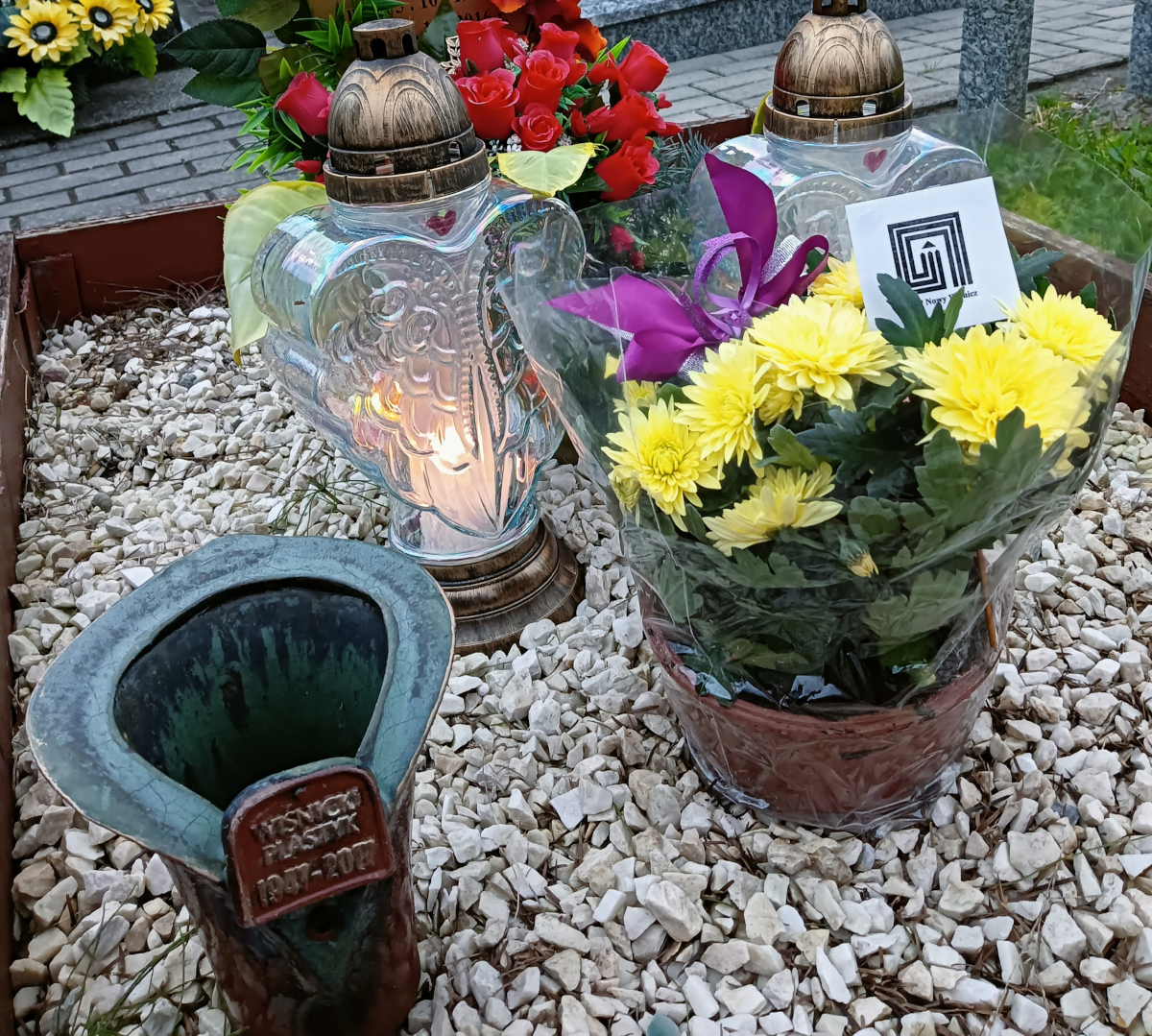 fotografia barwna przedstawia kwiaty oraz ceramiczne i szklane znicze na drewnianym nagrobku wyłożonym białym kamieniem