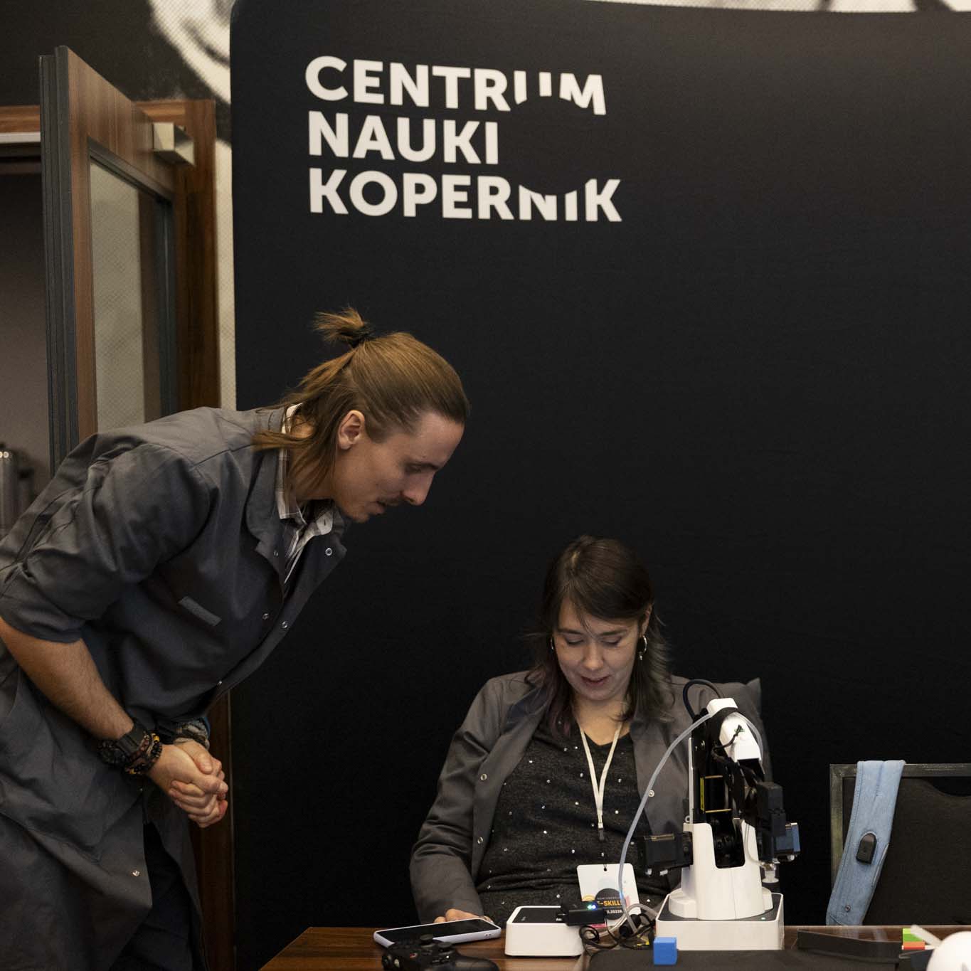 Na krześle siedzi kobieta a obok stoi mężczyzna nachylając się nad nią. Obydwoje mają szare fartuchy z logo Centrum Nauki Kopernik, za nimi stoi ścianka Centrum Nauki Kopernik. Na pierwszym planie znajduje się robotyczne ramię. 