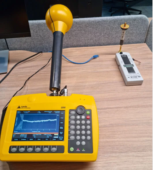 Zdjęcie przedstawia profesjonalny miernik firmy NARDA model SRM3006 leżący na stole obok amatorskiego miernika HFE35C.
