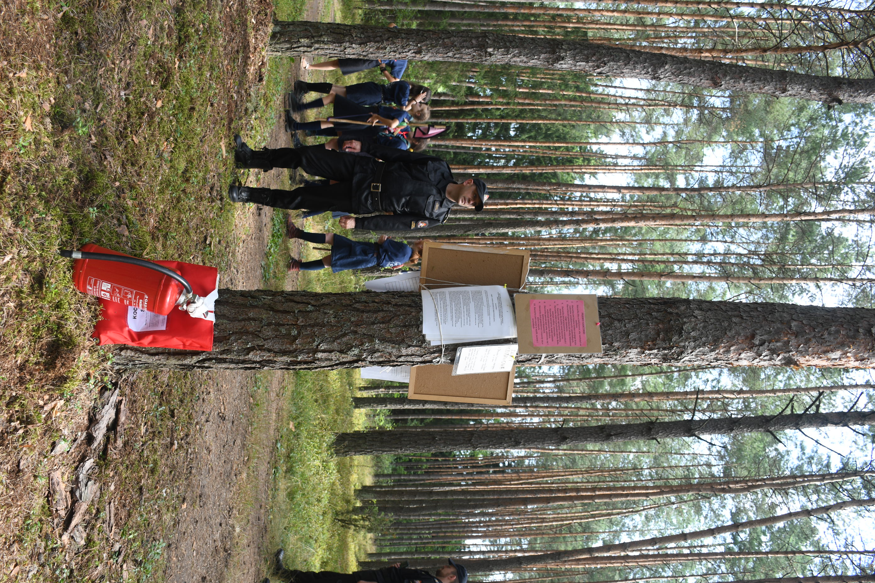 Strażak w lesie, za nim uczestnicy obozu, przy drzewie ustawiona gaśnica i koc gaśniczy