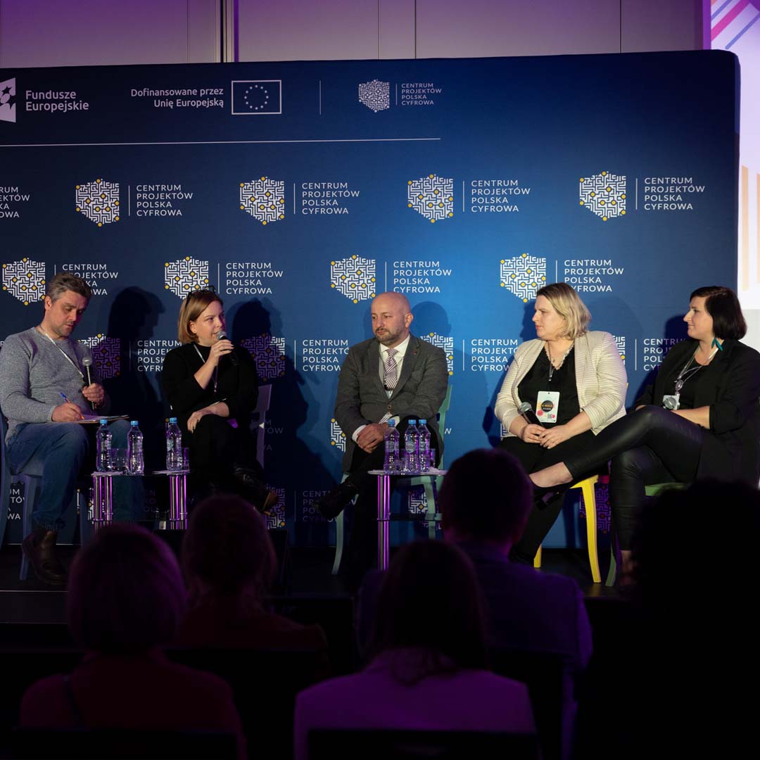 Pięciu panelistów siedzi na krzesłach na podium. Pani Oktawia Gorzeńska mówi do mikrofonu, reszta uczestników słucha wypowiedzi. Doktor Iwo Zmyślony robi notatki.