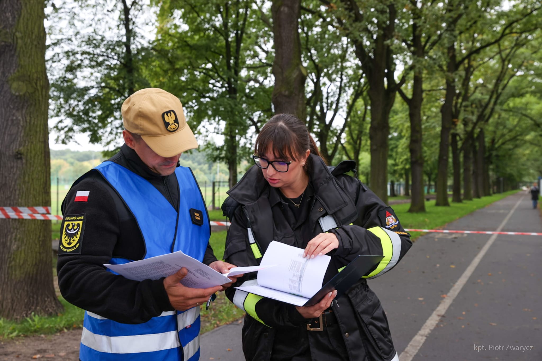 Dwóch strażaków (mężczyzna i kobieta) rozmawiają czytając dokumenty podczas ćwiczeń, za nimi widoczna droga i drzewa na poboczu