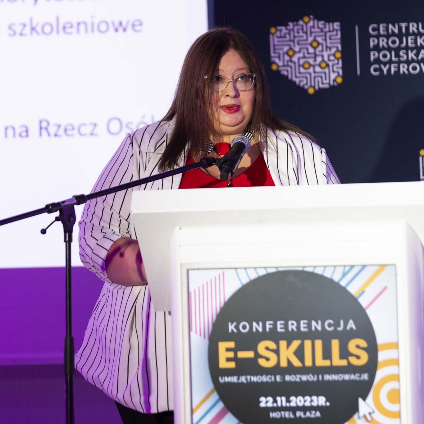 Przy mównicy stoi Pani Magdalena Janczewska. W ręku trzyma mikrofon patrząc na siedzących przed nią uczestników konferencji.