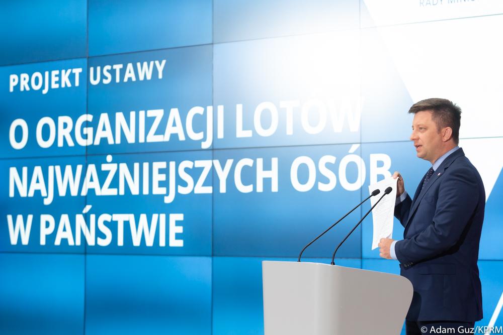Minister Michał Dworczyk mówi do mikrofonu na tle ścianki wizyjnej.
