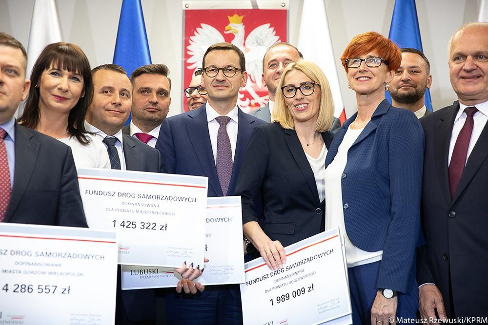 Premier Mateusz Morawiecki stoi wraz z samorządowcami, którzy trzymają w dłoniach czeki z dofinansowaniem.