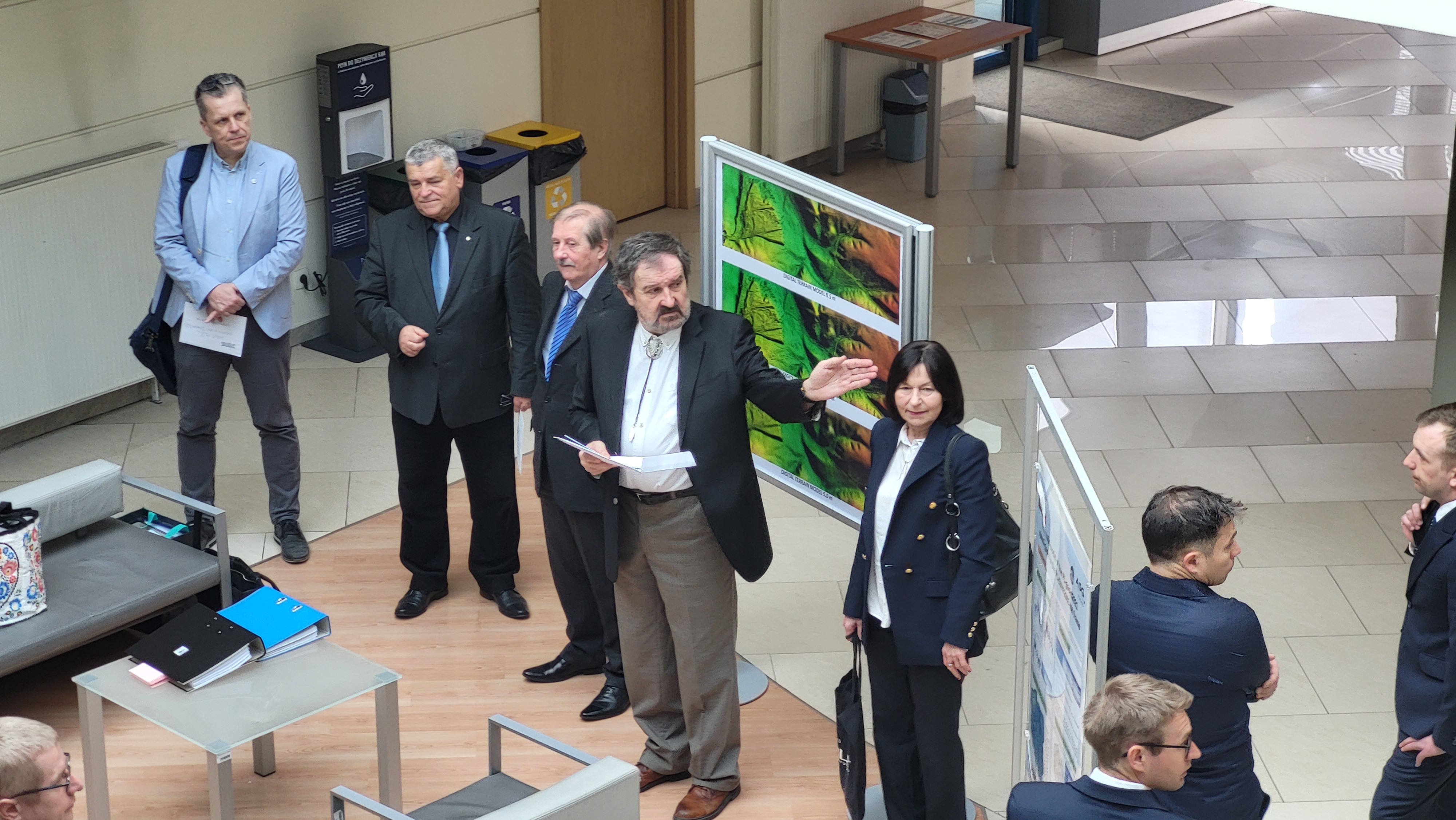 Zdjęcie przedstawia hol w siedzibie GUGiK przy ul. Olbrachta 94B z osobami przystępującymi do egzaminu słuchającymi wyjaśnień członków Komisji Egzaminacyjnej. 