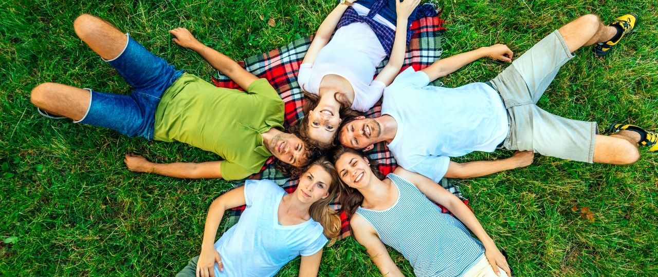 Na obrazku widać pięć osób leżących na trawie. Osoby ułożyły się w kółko, tak że ich głowy są blisko siebie, a nogi skierowane na zewnątrz. Leżą na kocu w kratkę. Trawa jest równa i zadbana.