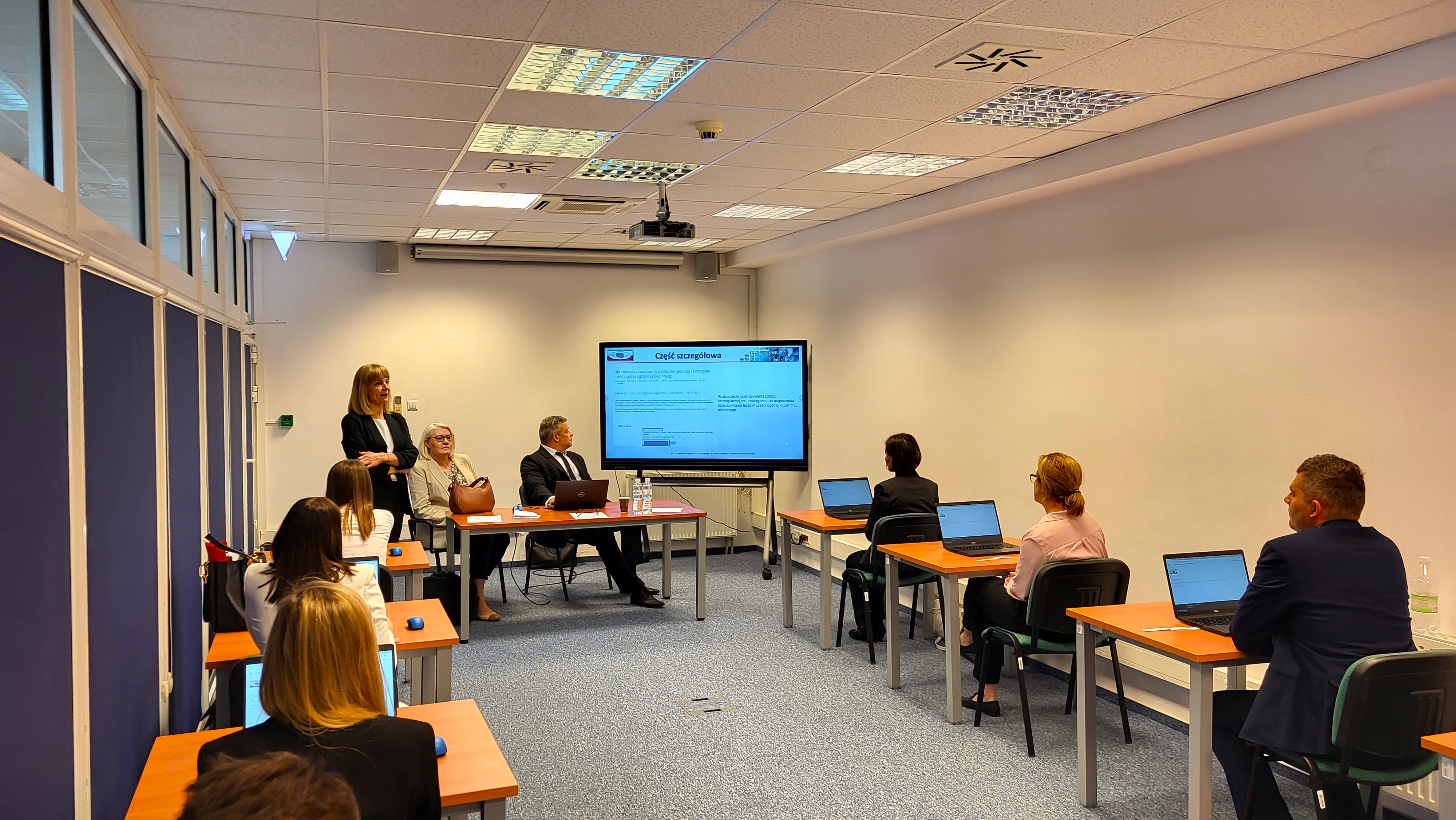  Zdjęcie przedstawia salę egzaminacyjną z członkami Komisji egzaminacyjnej i osobami przystępującymi do egzaminu siedzącymi pojedynczo przed laptopami.