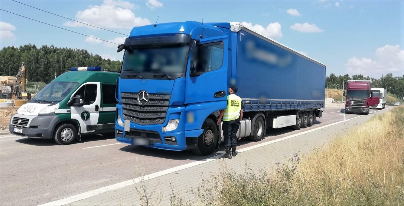 Inspektor stoi koło otwartych drzwi kabiny ciężarówki i pobiera dokumenty. Obok zestawu stoi radiowóz ITD.