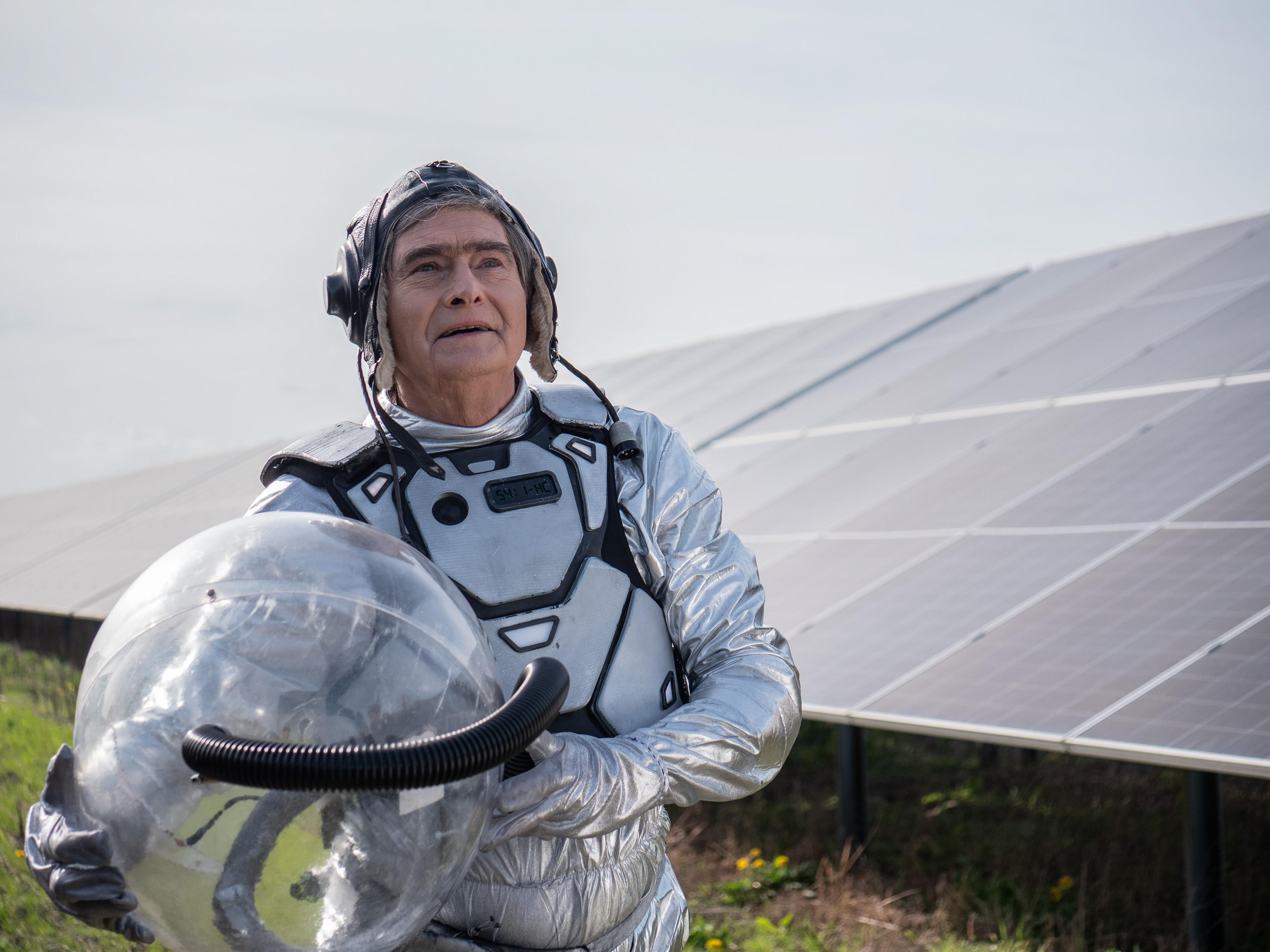 Uśmiechnięty mężczyzna w stroju astronauty patrzy w górę, w rękach trzyma hełm; obok niego rząd paneli fotowoltaicznych