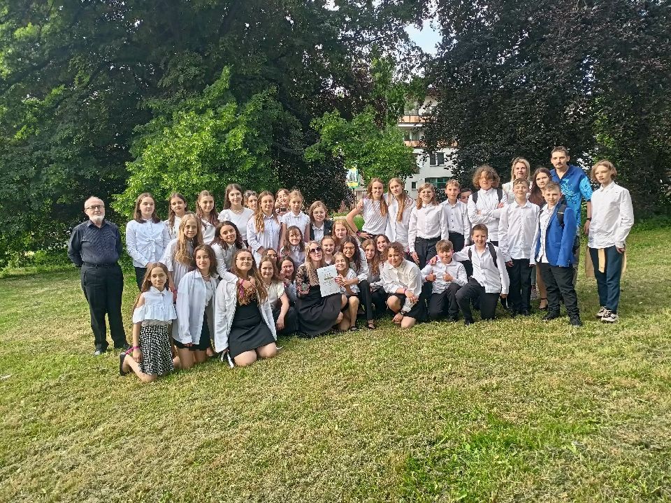 Zdjęcie. Zespół chórzystów wraz z nauczycielami pozują do zdjęcia na wolnym powietrzu na trawie.