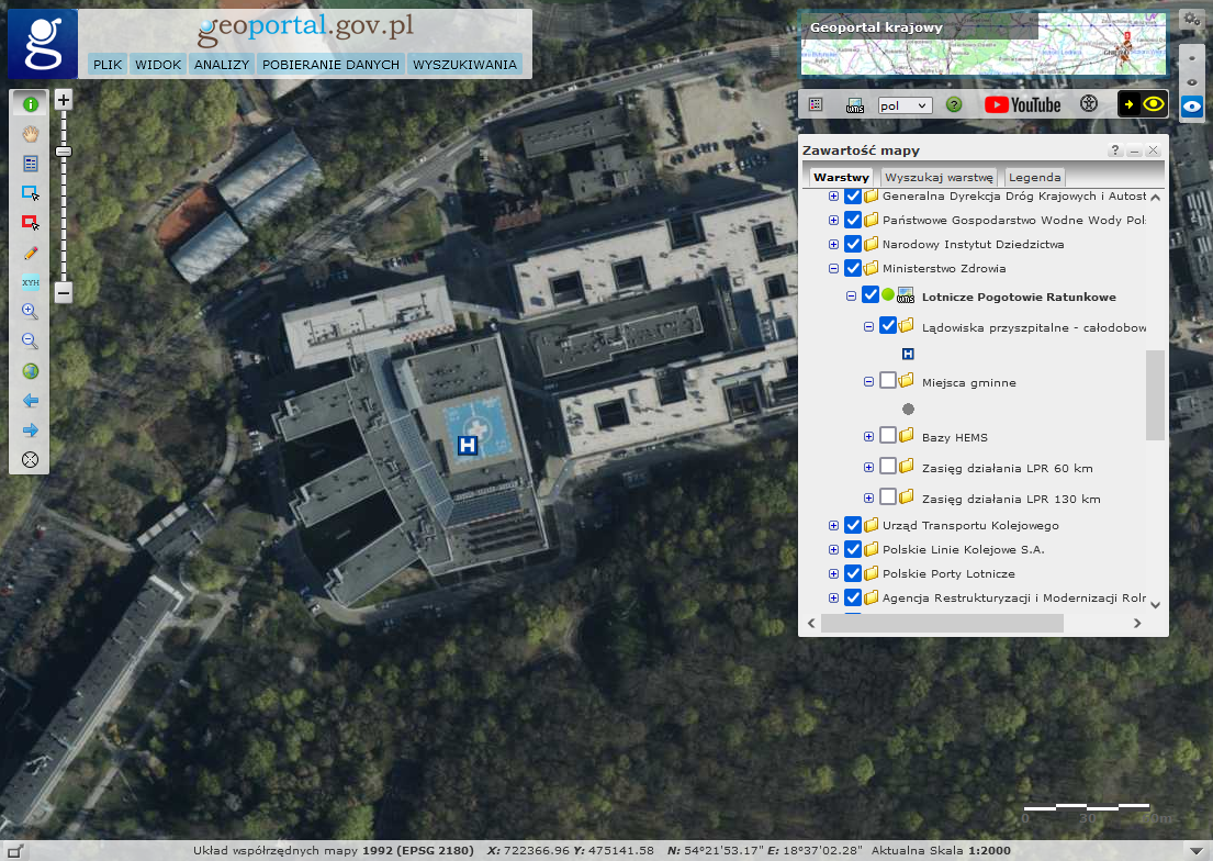 Ilustracja przestawia widok Geoportalu z przyblizeniem do jednego lądowisk przyszpitalnych. Na mapie podkładowej znajduje się ortofotomapa z widokiem szpitala i infrastruktura lądowiska.
