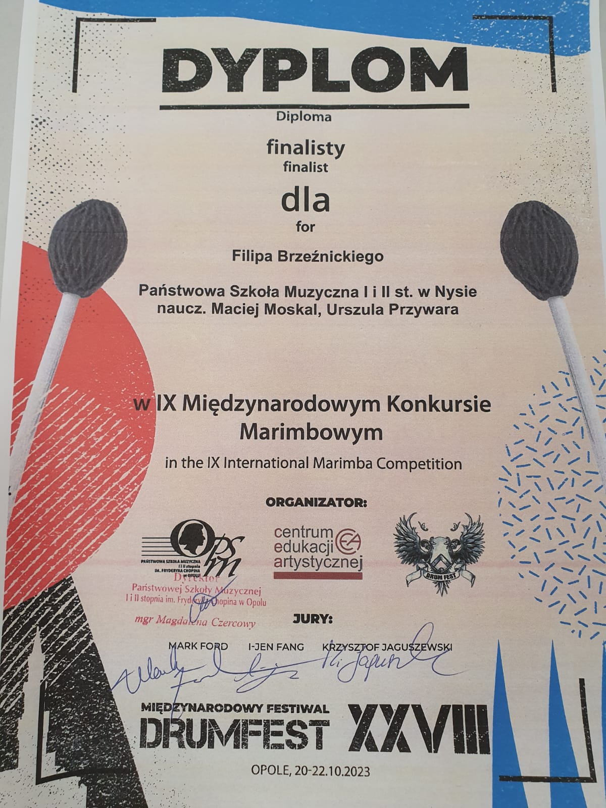 Dyplom na szarym papierze u góry fragment paska niebieskiego z boku czerwonego i czarnego sprawy i z lewej strony pałki do marimby od góry napis w kolorze czarnym dyplom kreska diploma finalisty finalisty dla Filipa Brzezińskiego państwowa szkoła muzyczna pierwszego i drugiego stopnia w Nysie nauczyciel Maciej Moskal Urszula przywara w 9 międzynarodowym konkursie marimbowym organizator logo szkoły z podpisem dyrektorki Centrum Edukacji Artystycznej i bliżej nieokreślony Orzeł bądź dwa od niżej podpisy jurorów Mark Ford i Jean-Fang Krzysztof Jaguszewski międzynarodowy festiwal drum Fest 18 Opole 20 22 października 2023