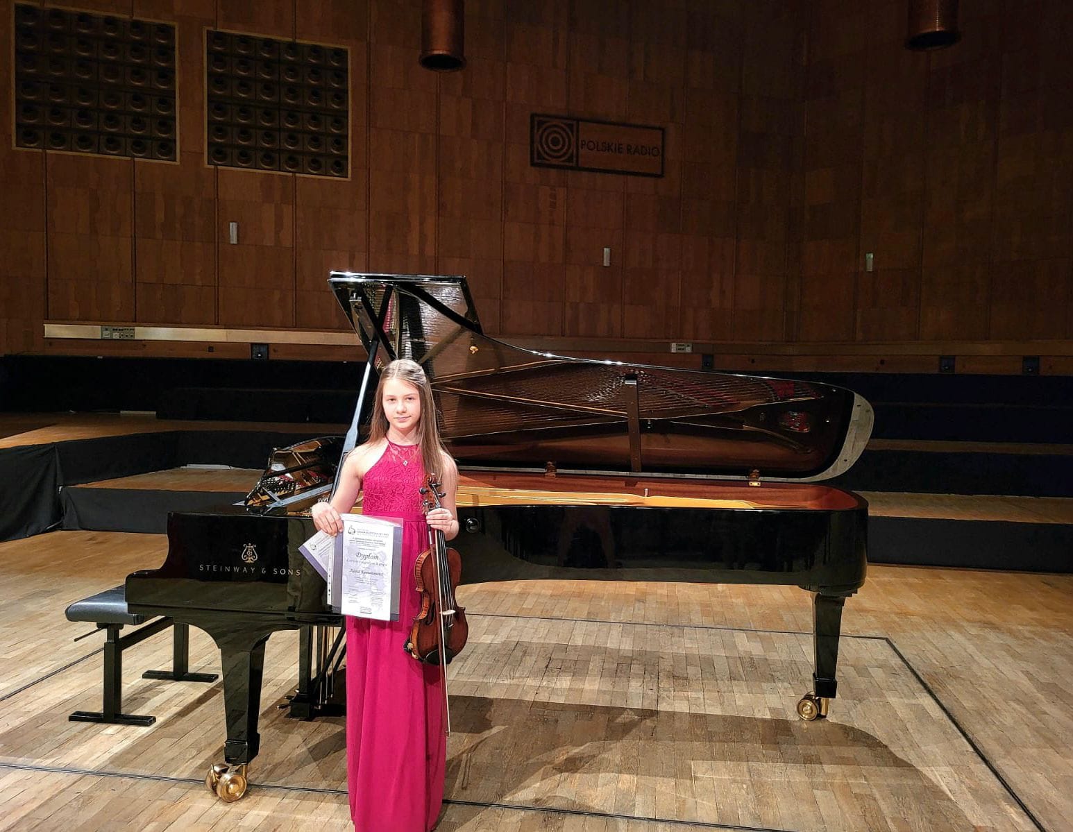 Na zdjęciu widoczna uśmiechnięta dziewczyna w bordowej sukience trzymająca w prawej ręce dyplomy, w lewej skrzypce. W tle widoczny czarny fortepian. 