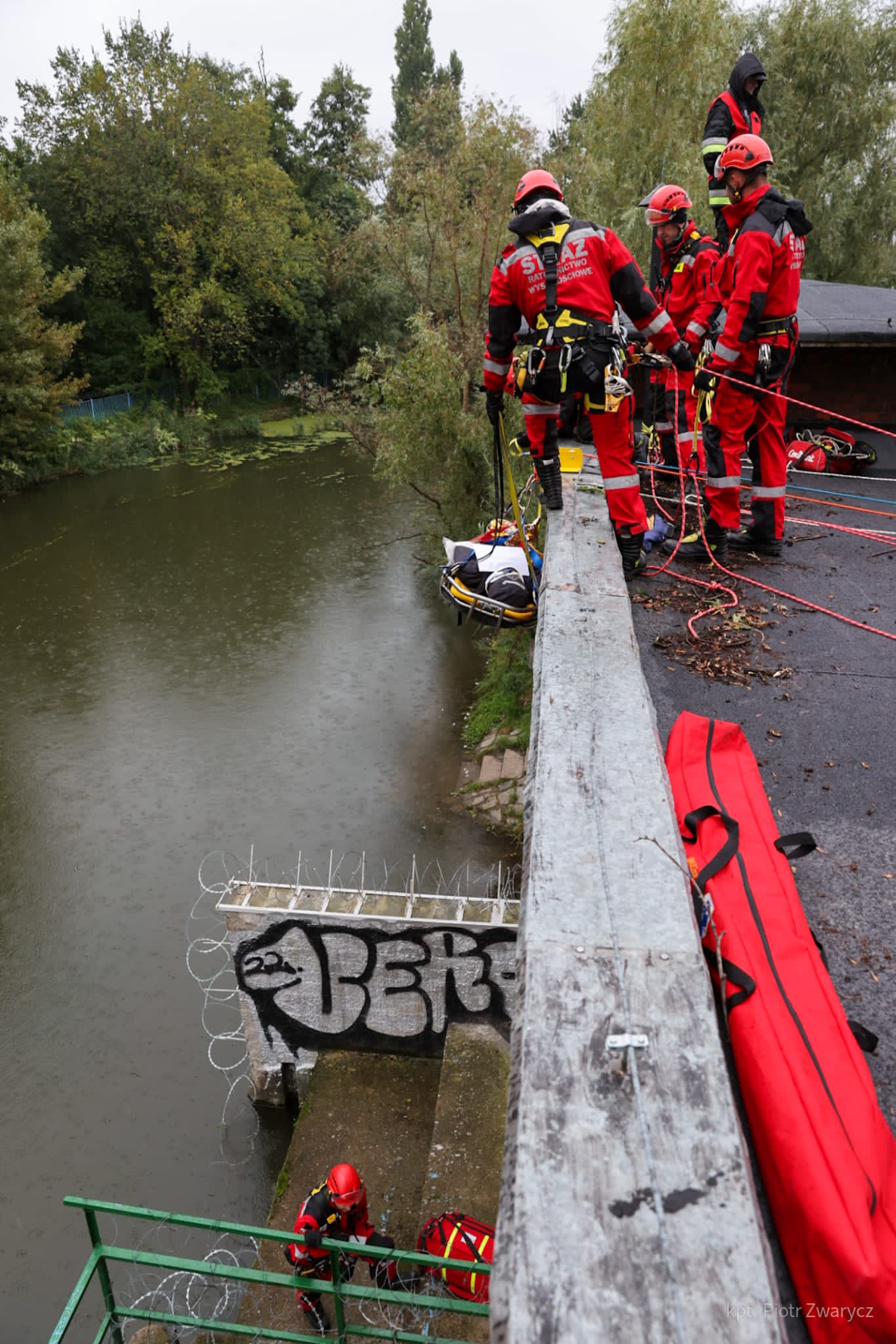Zapora na rzece, na której widoczni są strażacy ze specjalistycznej grupy ratownictwa wysokościowego w umundurowaniu w kolorze czerwonym.