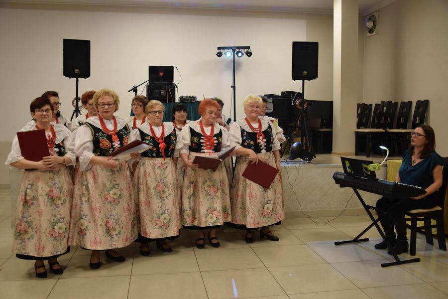 Ubrane w stroje ludowe kobiety stoją w dwóch rządach podczas występu. Obok inna kobieta gra na organach